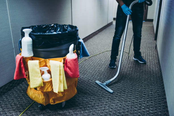 Janitor vacuuming the office corridor carpet, with cleaning products and wipes by the garbage bin for disinfection.