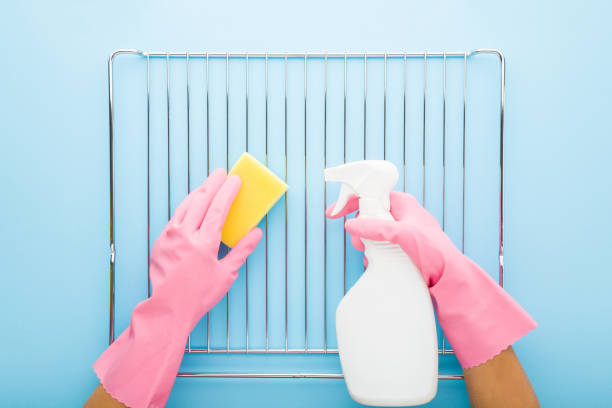 Woman hands in rubber protective gloves holding detergent bottle, sponge and washing steel grill grate for kitchen oven on light blue table background. Closeup. Point of view shot. Top down view.