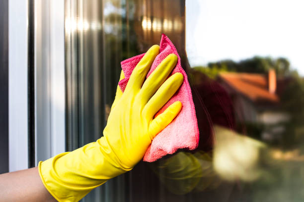 A hand in yellow protective glove cleaning the glass window pane with a pink rag.