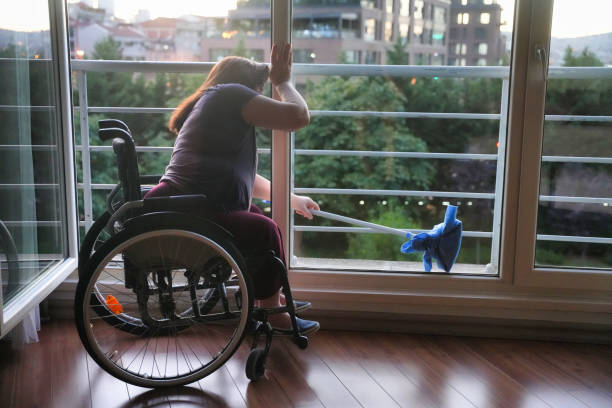 A woman in a wheelchair reaches outside with a mop to clean the exterior of a glass balcony door.