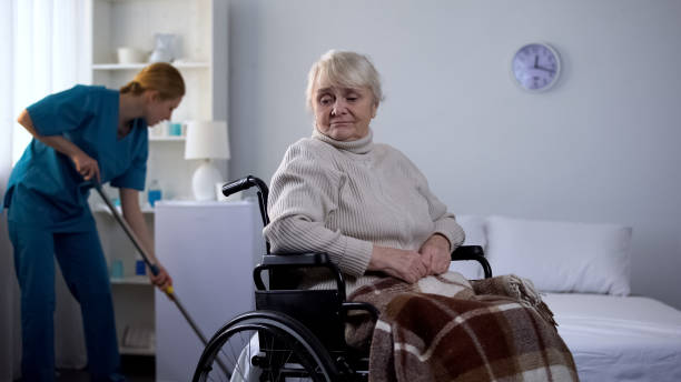 Sad woman in wheelchair watching hospital janitor cleaning room, medical center.