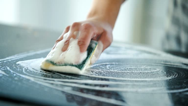 Closeup of unrecognizable woman cleaning glass ceramic stove top with a detergent and a soft cleaning sponge.