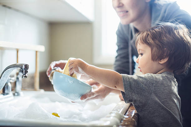 Toddler boy in blue diaper helps wash dishes, smiling mother supervises.