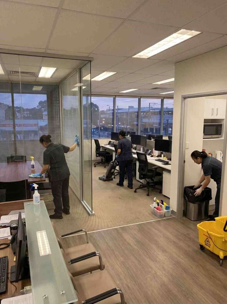 Professional cleaners vacuuming carpets, wiping glass partitions and emptying bins during after-hours office cleaning in a Burwood, NSW workplace.